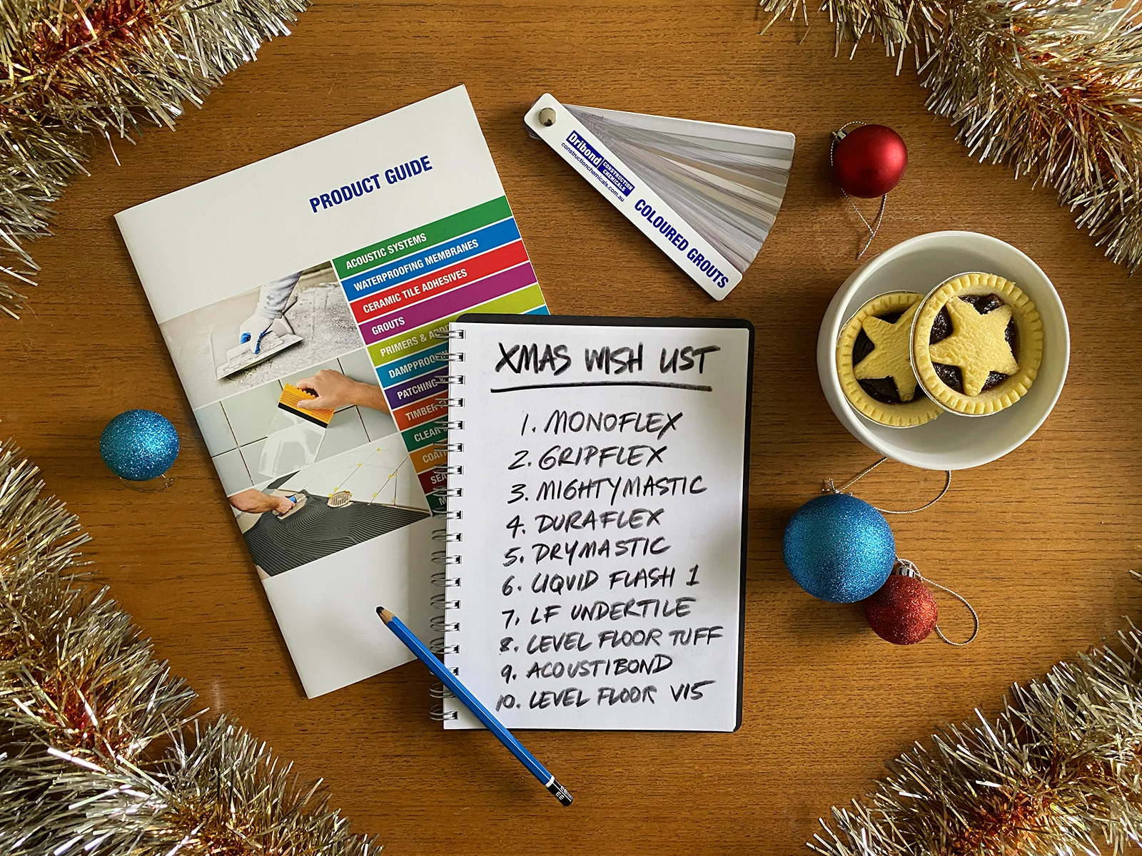 Dribond Xmas Wishlist Hand-written "Xmas Wish List", a Dribond Construction Chemicals product guide and a grout colour chart on a wooden table top, surrounded by tinsel, baubles and mince pies.